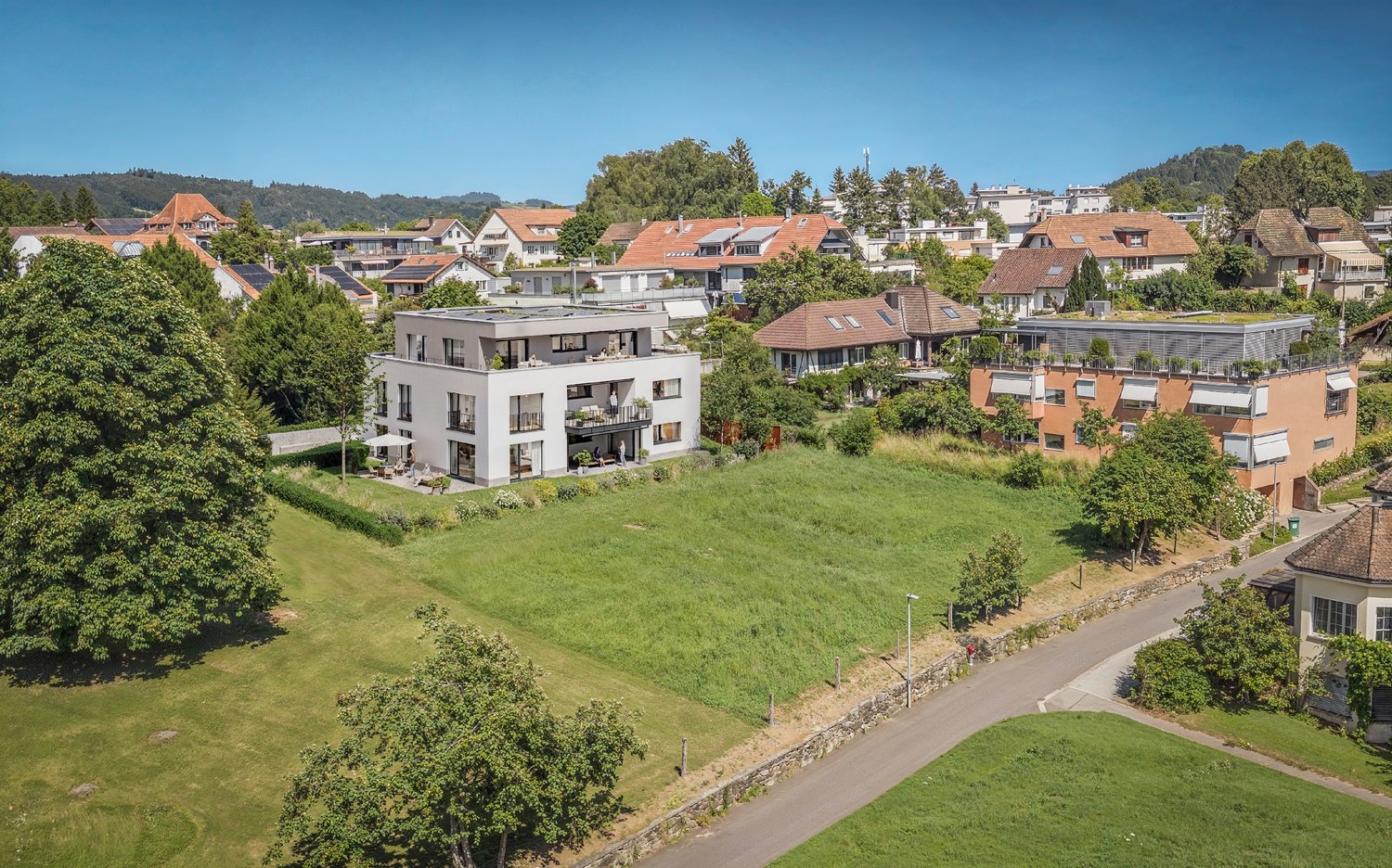 Modern apartment building with balconies, located in a residential area with greenery and mountains in the background.