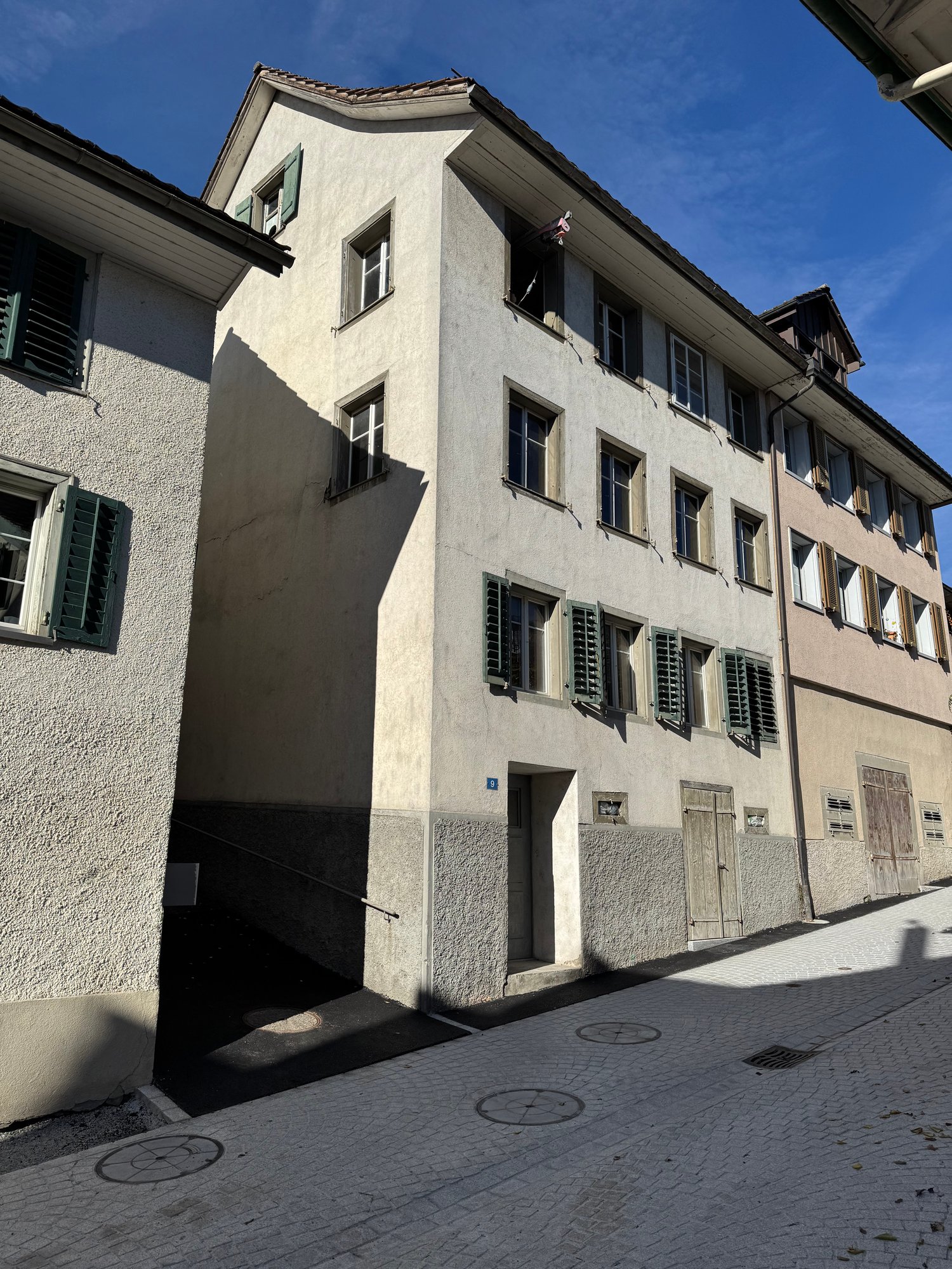 Concrete building with multiple windows and green shutters, street entrance, paved street.