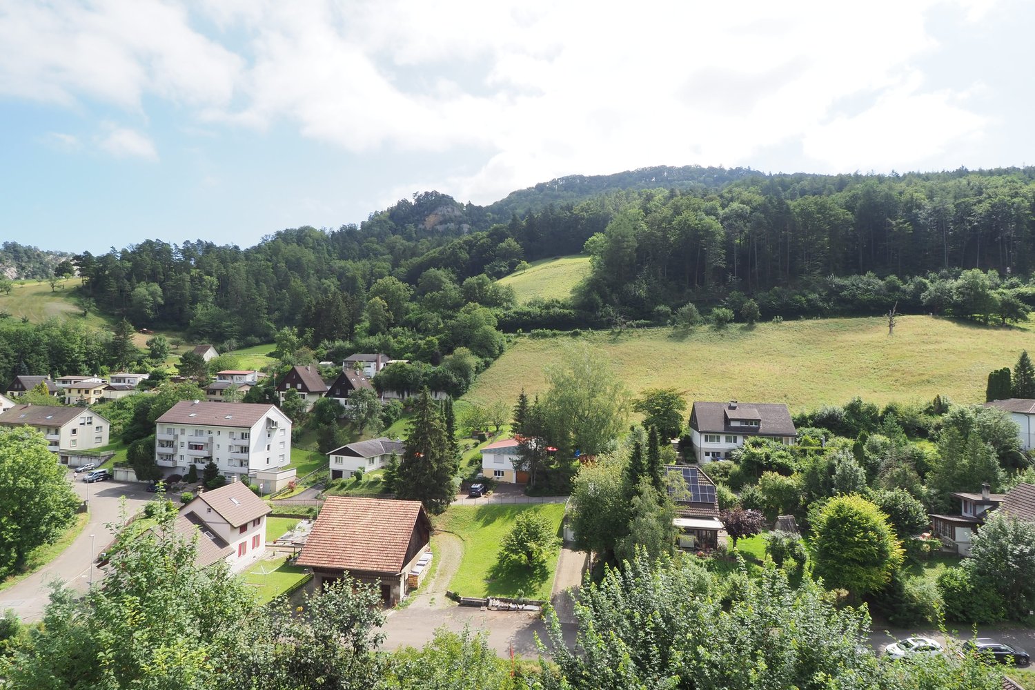 The image shows a scenic rural landscape with a village nestled in a valley surrounded by lush green hills and forests. The houses have red tile roofs and are surrounded by trees and vegetation. There are grassy fields and a winding road visible in the fo