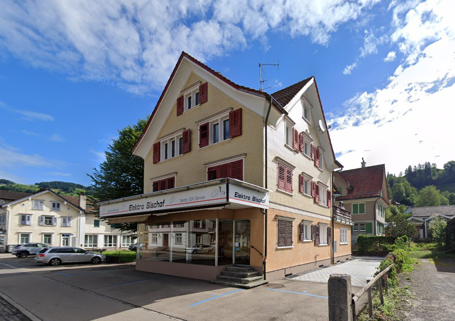 3-story building, brick facade, red shutters, storefront at ground level, small car park in front, green trees, neighboring buildings visible in background