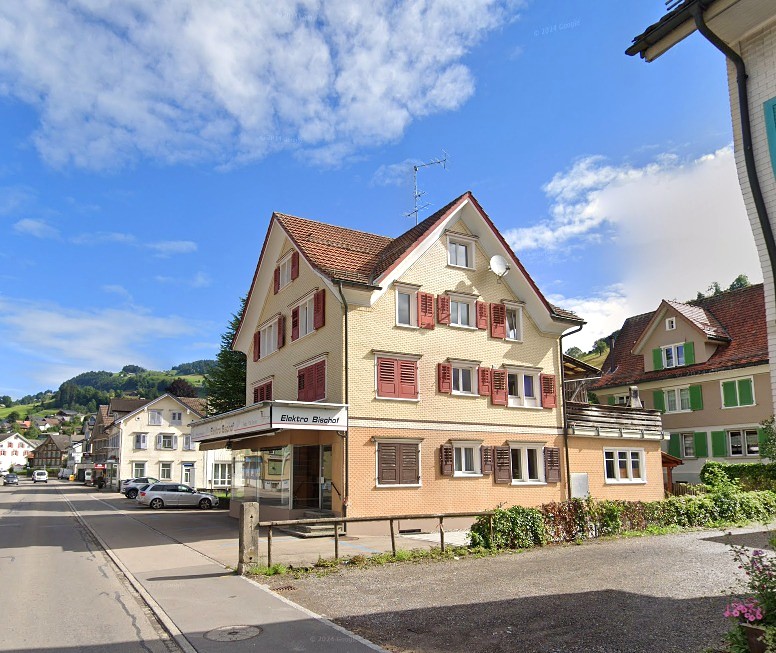 A large building on the street with a sign 'Elektro Bischoff', many windows, red shutters, with cars parked in front