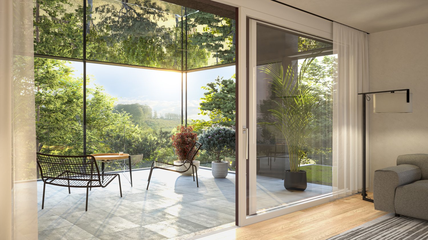 Large glass windows overlooking a lush, green outdoor area with trees and plants. Sliding glass doors lead to a balcony or terrace. The room has a modern, minimalist design with a couch, chairs, and a small table.