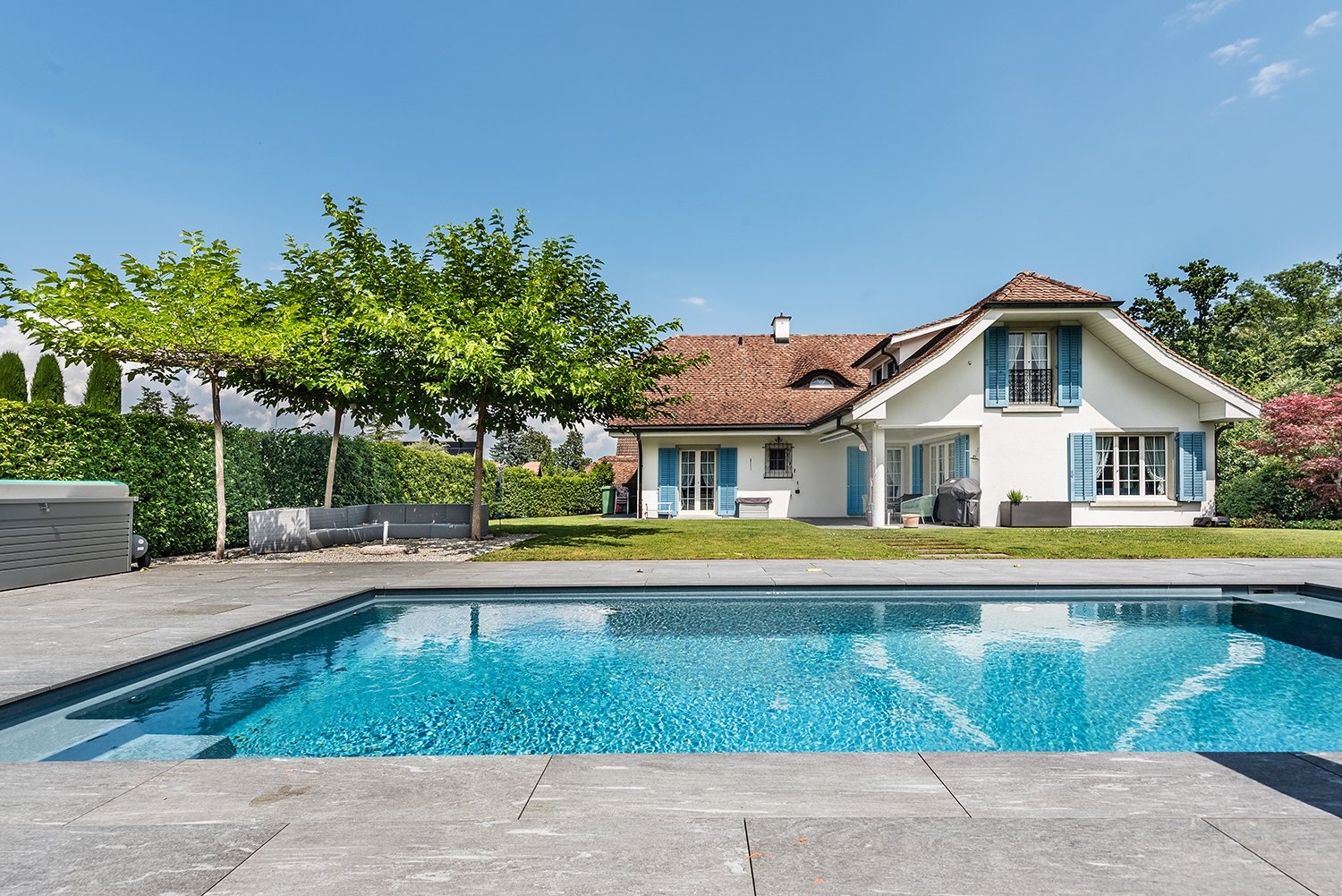 2 story white house, brown roof, pool, lush green lawn, hedges, gravel driveway, trees