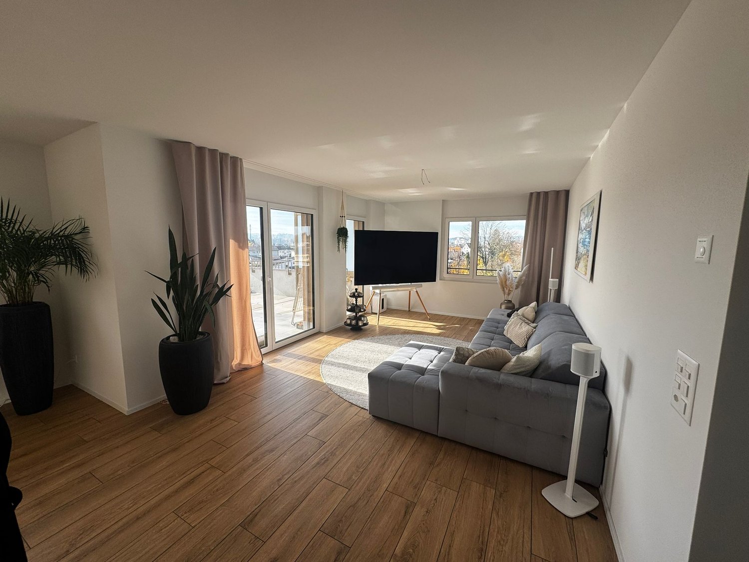 Open living area with L-shaped sofa, large TV, floor-to-ceiling windows, balcony with city view