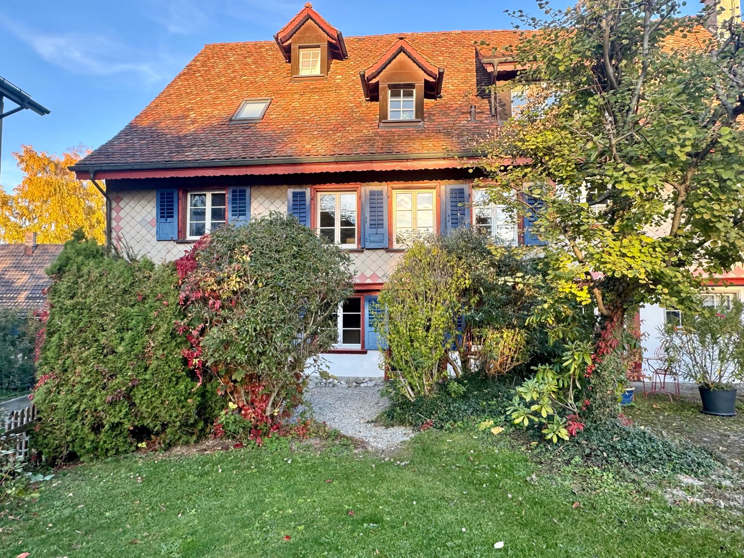 Red tiled roof, white exterior walls, blue window shutters, multiple windows, shrubs, pathway, gravel