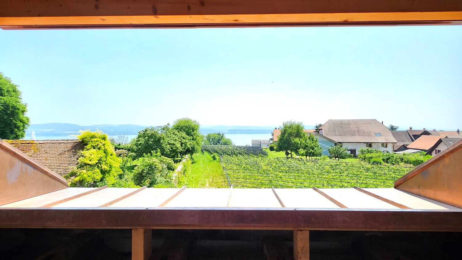 The image shows a scenic view from a balcony or terrace. The view includes a lush green vineyard or orchard, with houses and buildings in the distance. The sky is a bright blue color, and the landscape appears to be in a rural or semi-rural area.