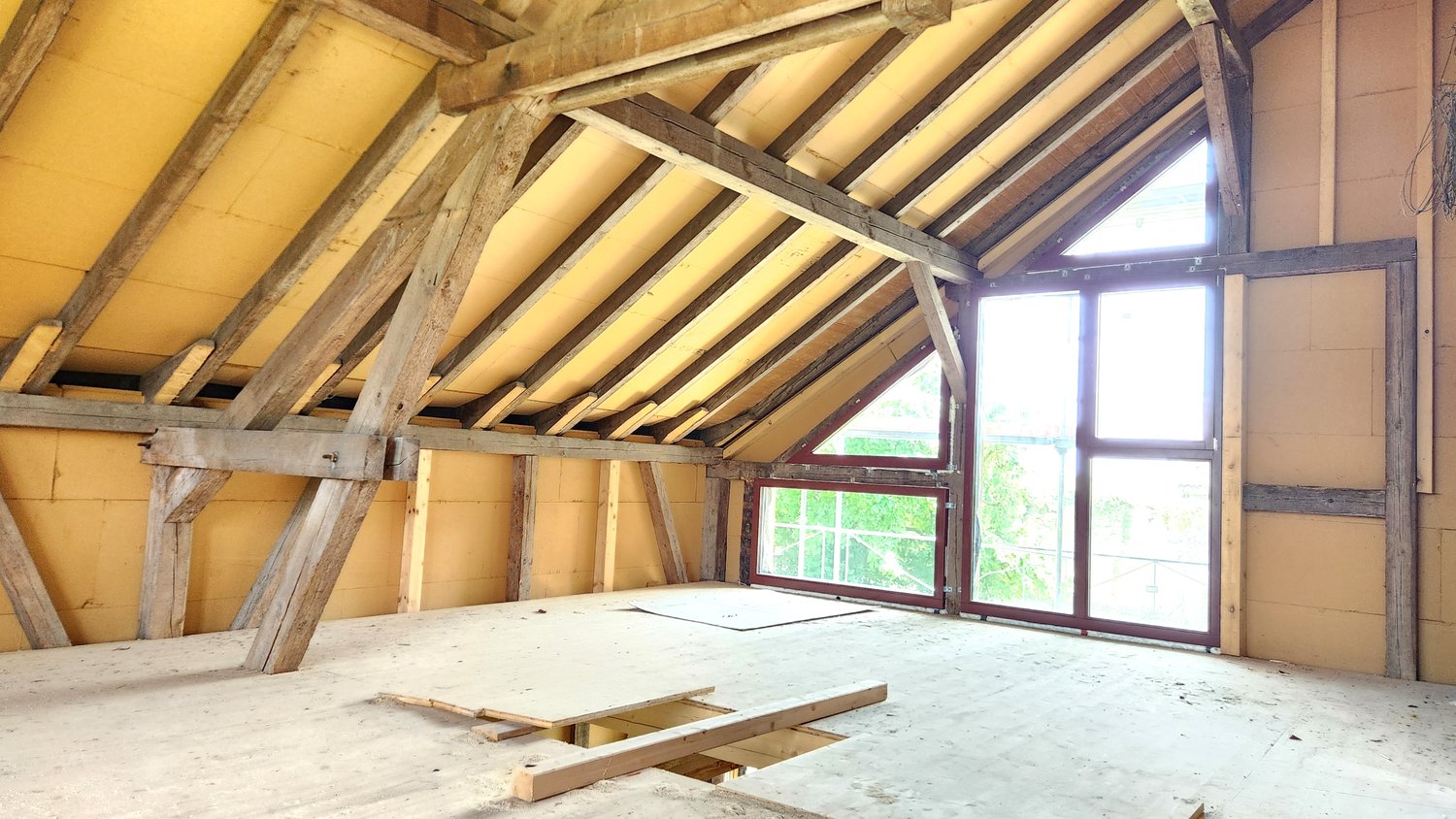 The image shows the interior of an attic space with exposed wooden beams and trusses supporting the roof structure. The attic appears to be in the process of renovation or construction, with some construction materials visible on the floor. Large windows