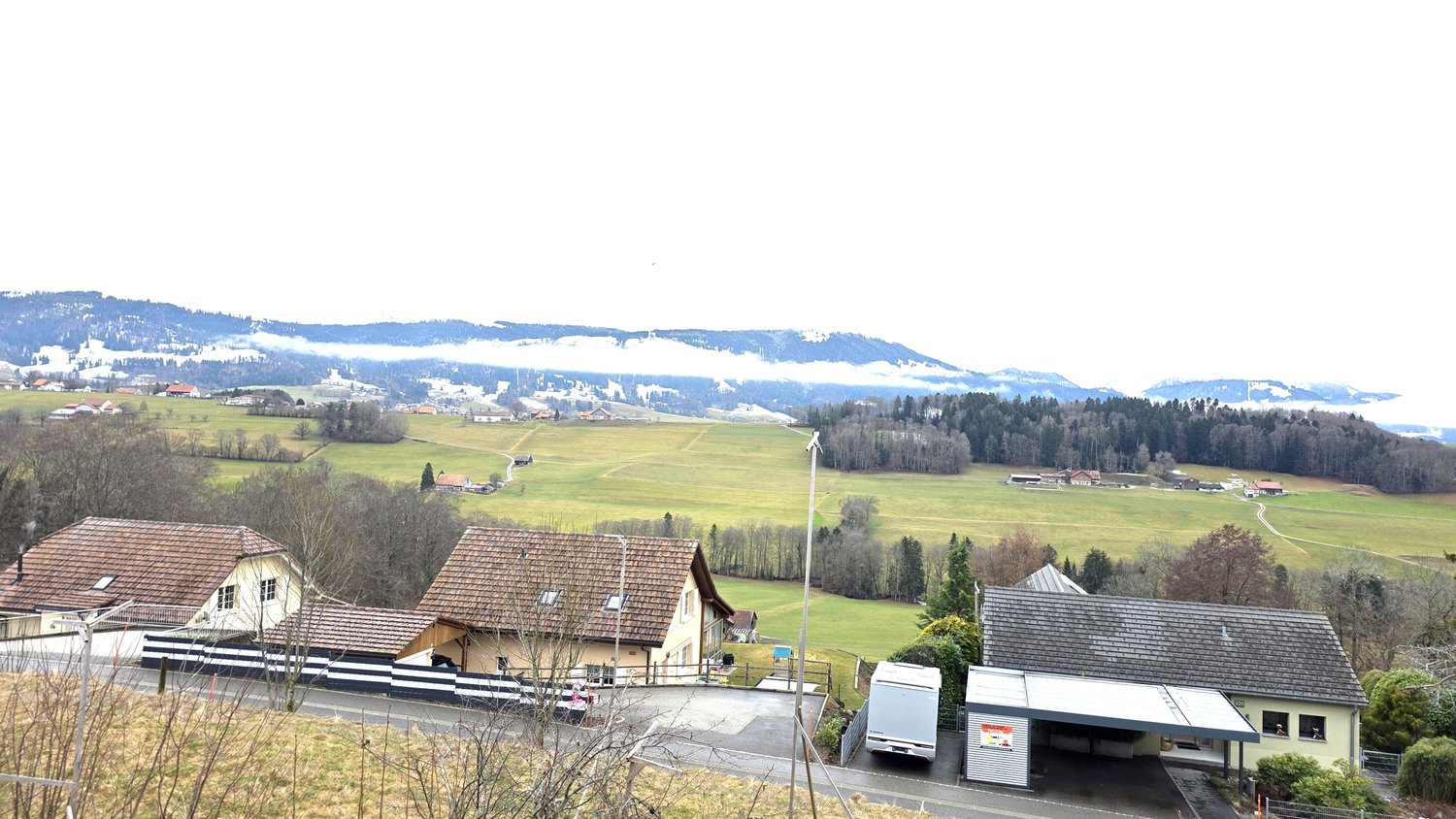Aerial view of a village with several houses, wide green field, trees, hills, and a cloudy sky