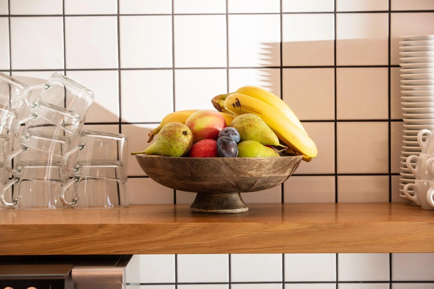 Light wooden countertop, bowl of fruits, stack of cups, tiled backsplash, oven
