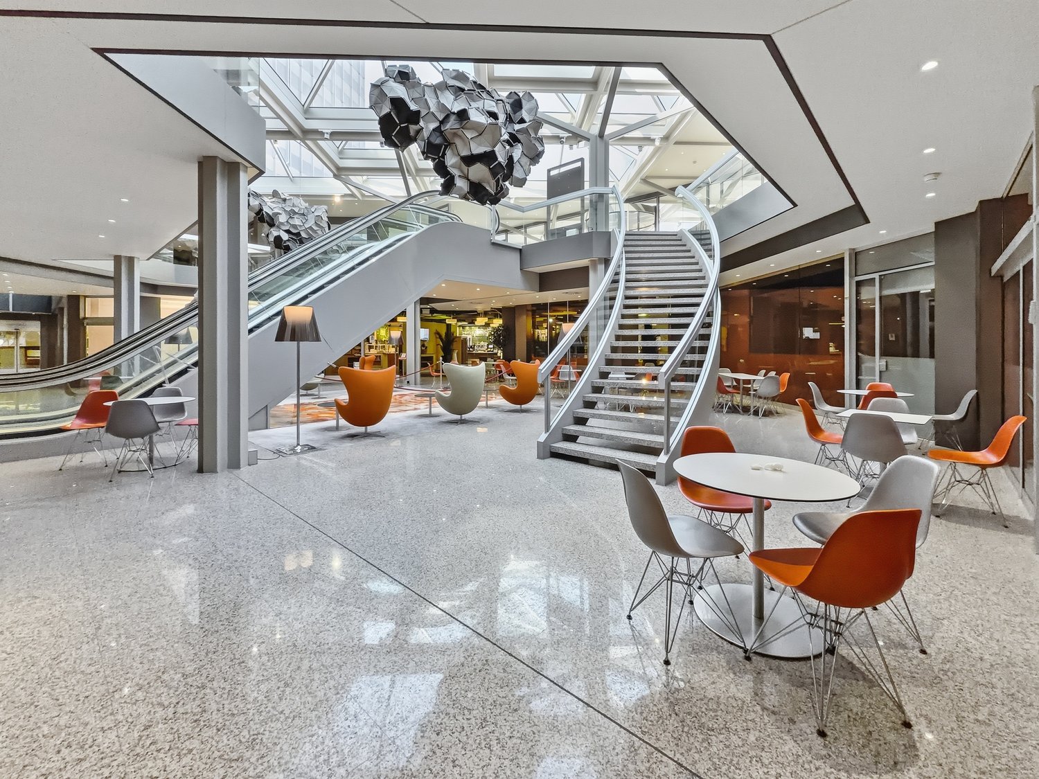 modern interior with white and orange chairs, white tables, large staircase, escalator, glass ceiling