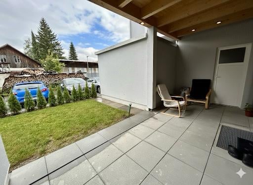 A modern house exterior with a patio area featuring tile flooring, two white outdoor chairs, a wooden canopy, and a small garden with plants. Cars are parked along the side of the house, and a building is visible in the background.