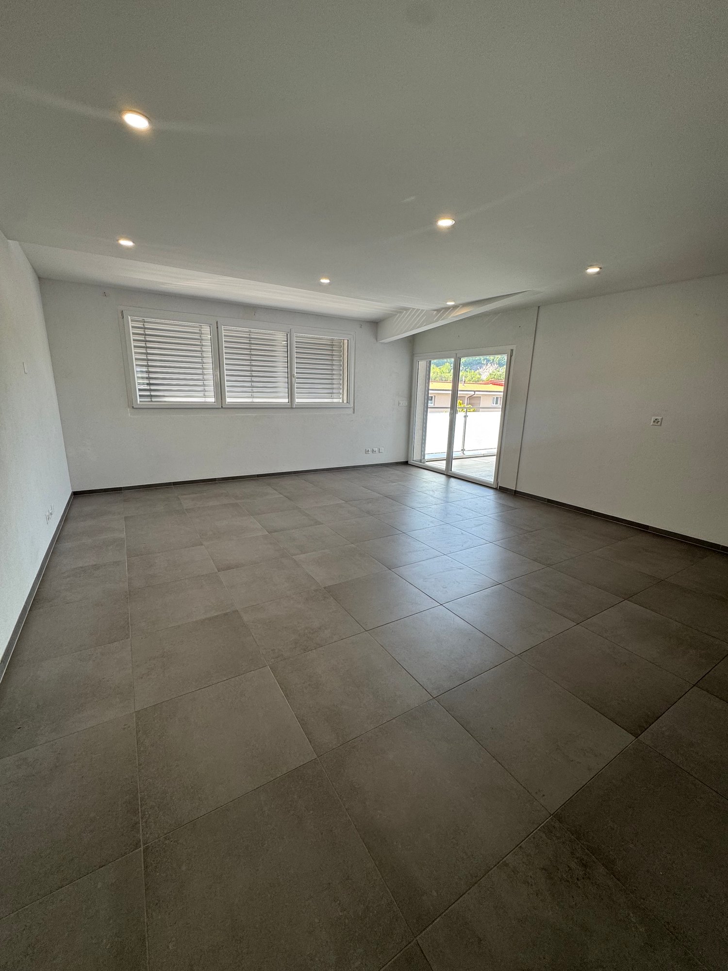 Large open living space with gray tile flooring, white walls, recessed lighting, and sliding glass doors leading to an outdoor area.