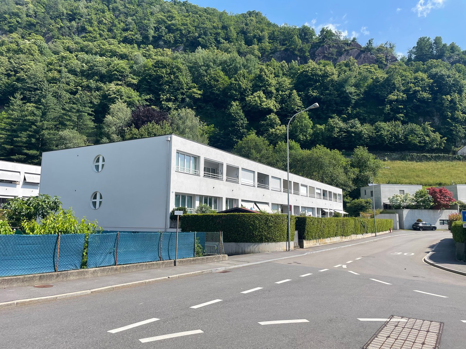 white apartment building, 3 stories, street, greenery, mountain, car parked in front
