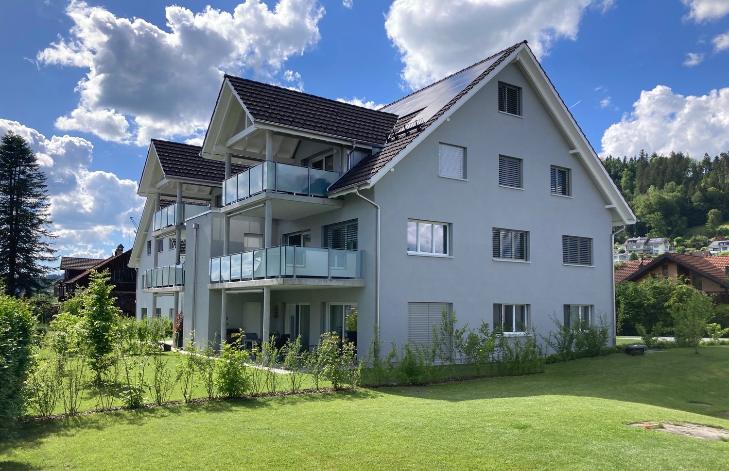 A two-story building with a brown roof, solar panels, a glass balcony on the second floor, and a garden in front.