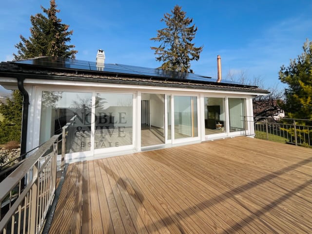 Single-story house with a large wooden deck or patio, surrounded by trees. The house has large windows and a chimney, suggesting it may have a fireplace. Solar panels are visible on the roof.