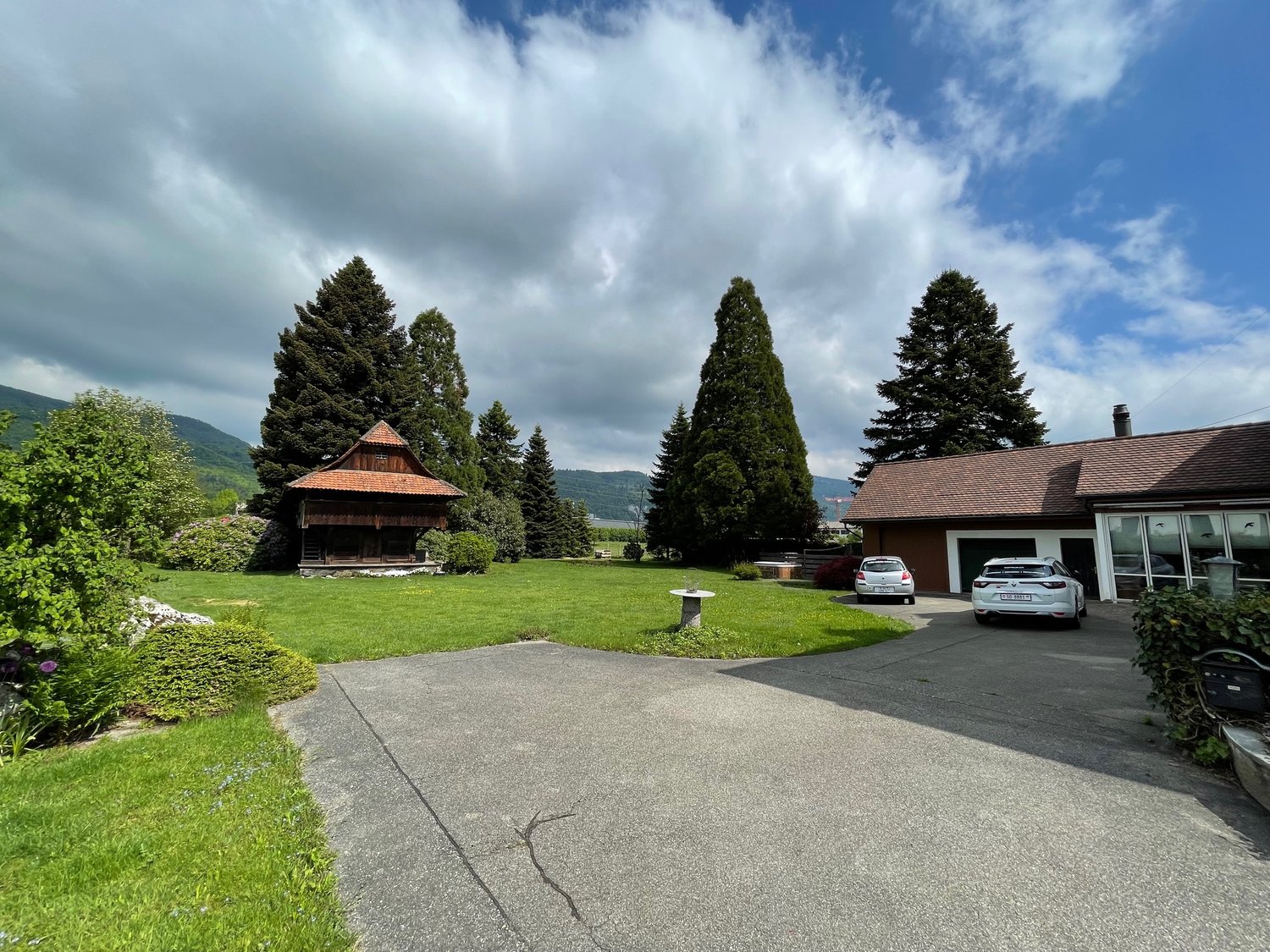 House exterior, parking area, two cars parked, wooden barn, lush green lawn, trees, mountainous background