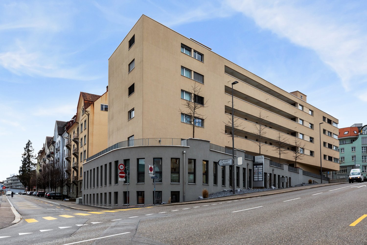 Modern multi-story building with beige exterior, glass windows, and a curved section at the base.