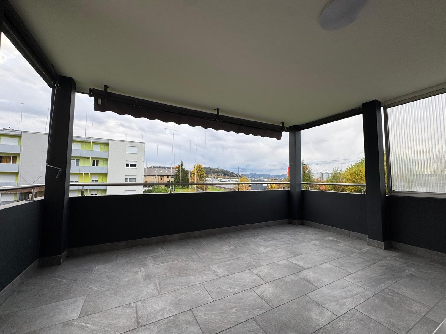 Large balcony, grey tiles, glass windows, metal railings, clear sky, distant buildings