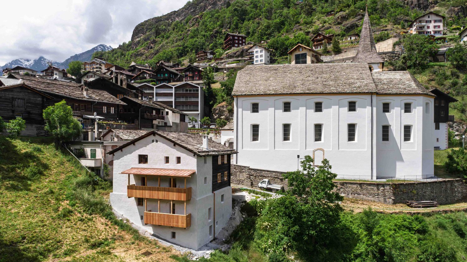 The image shows a picturesque alpine village nestled in a mountainous landscape. The buildings have traditional Swiss architectural styles, with wooden balconies and sloped roofs. There are several multi-story buildings, including a large white building w