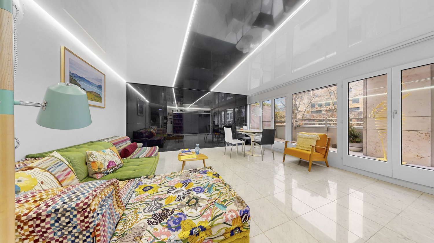 Spacious and modern living room with colorful and patterned furniture, including a large green sofa, a patterned armchair, and a coffee table. The room has large windows providing natural light, and the walls feature artwork and lighting fixtures. The flo