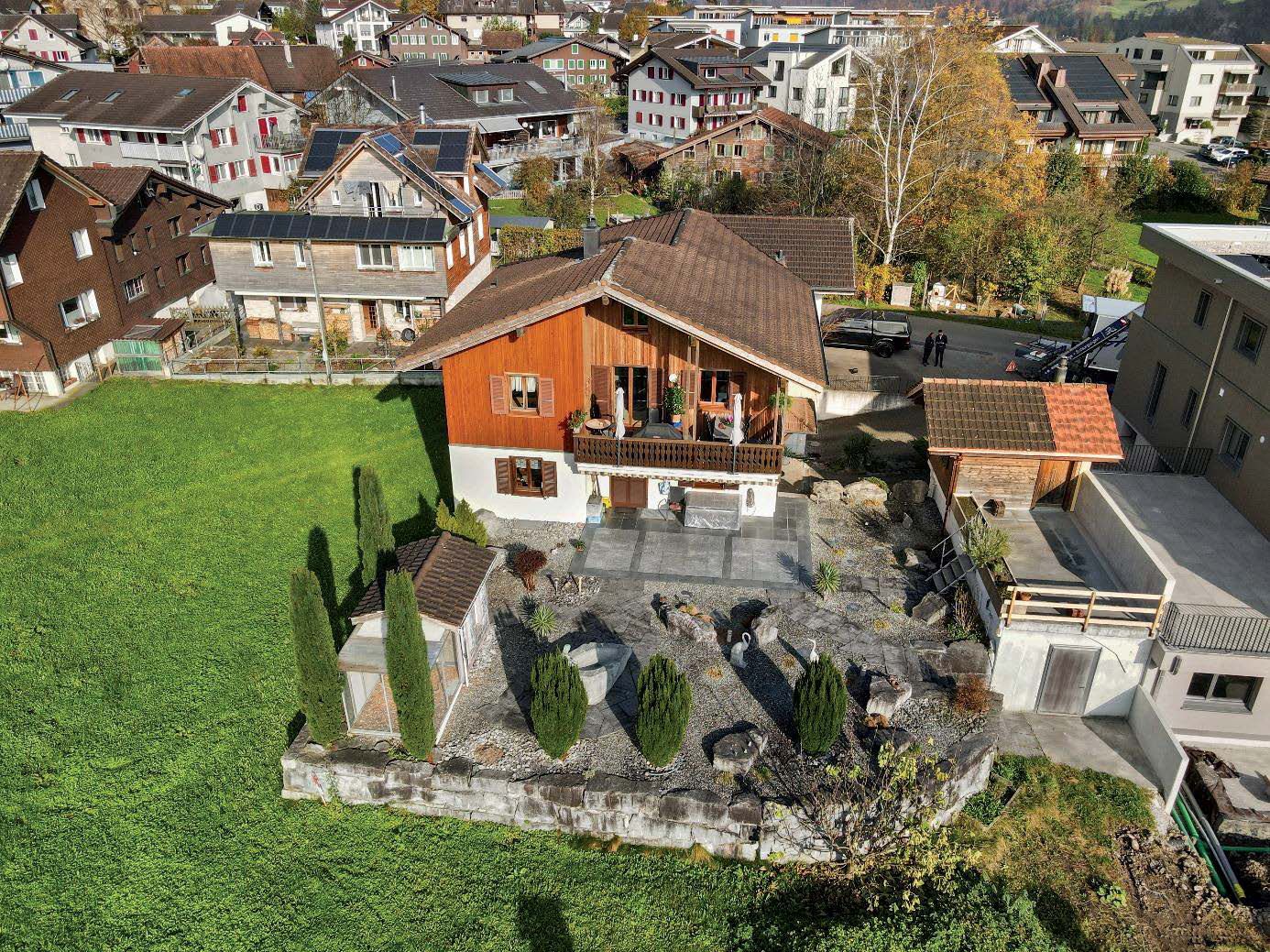Two-story wooden chalet-style house with a balcony, surrounded by a landscaped garden with trees and shrubs. The house is situated in a residential area with other houses and buildings visible in the background.