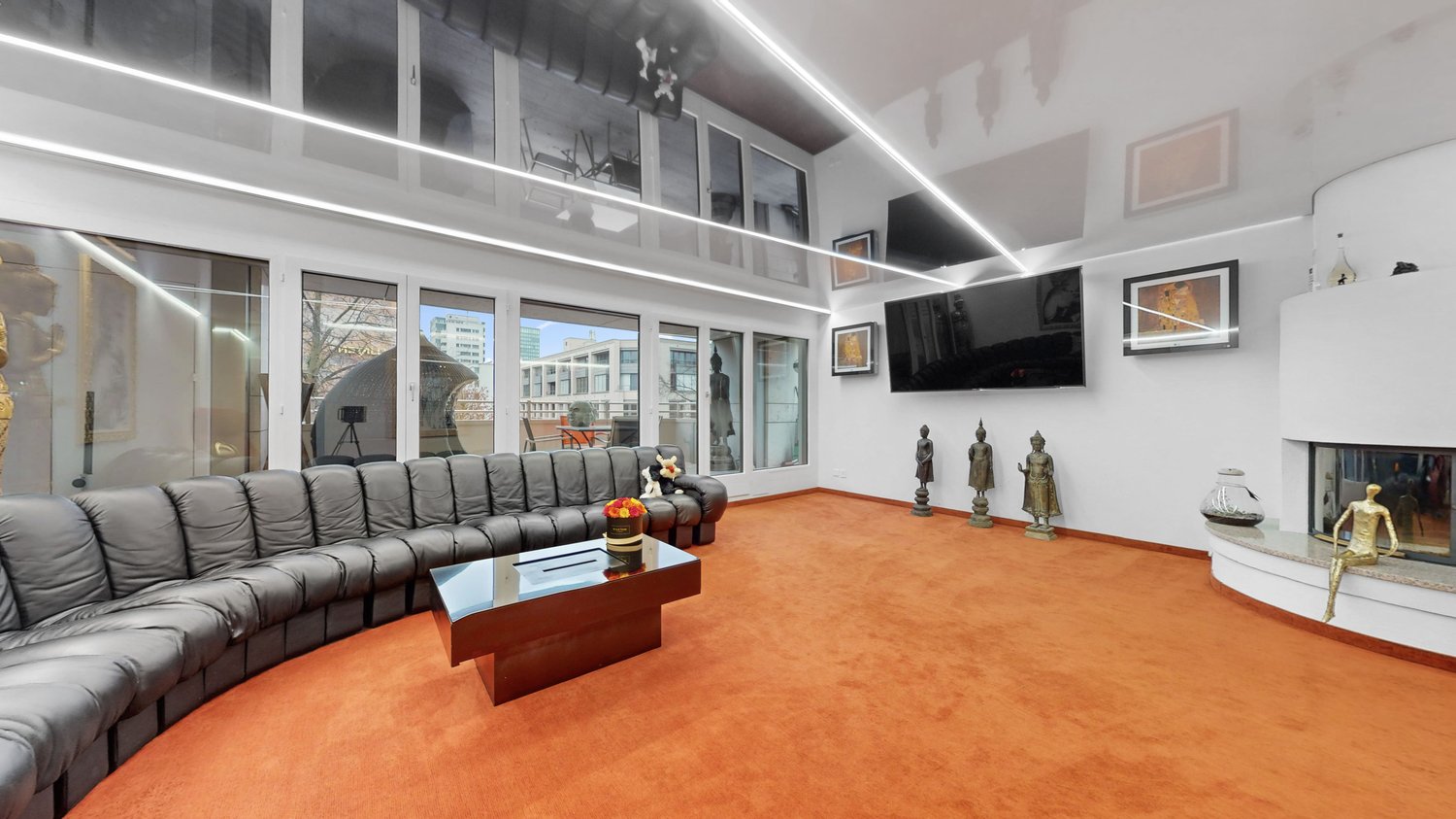 Spacious living room with large windows, black leather sectional sofa, wooden coffee table, and decorative statues. The room has a modern and minimalist design with high ceilings and track lighting.