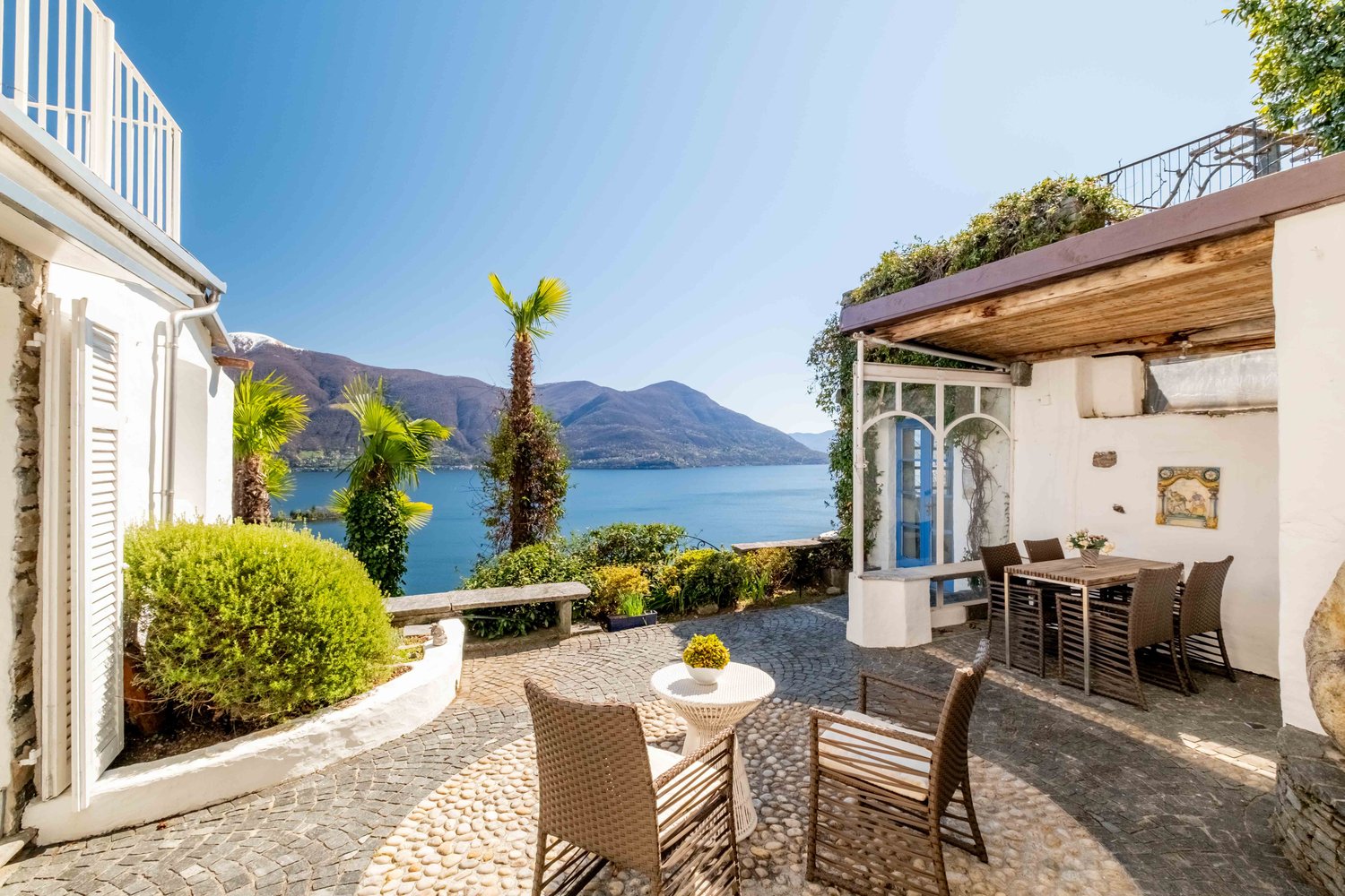 Outdoor patio with wicker furniture, overlooking a lake surrounded by mountains. The property has a white exterior with a wooden roof and palm trees in the garden.