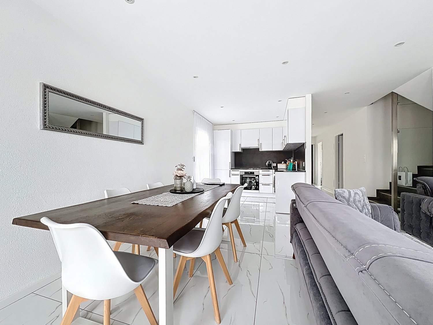 dining area with wooden table and white chairs, white modern kitchen, gray couch, white tile floor, mirror