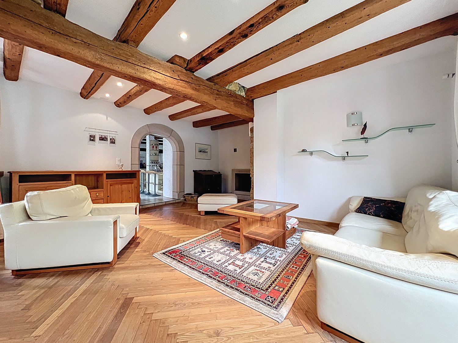 white walls, wooden ceiling, white leather sofa, ottoman, glass coffee table, rug, wooden floor