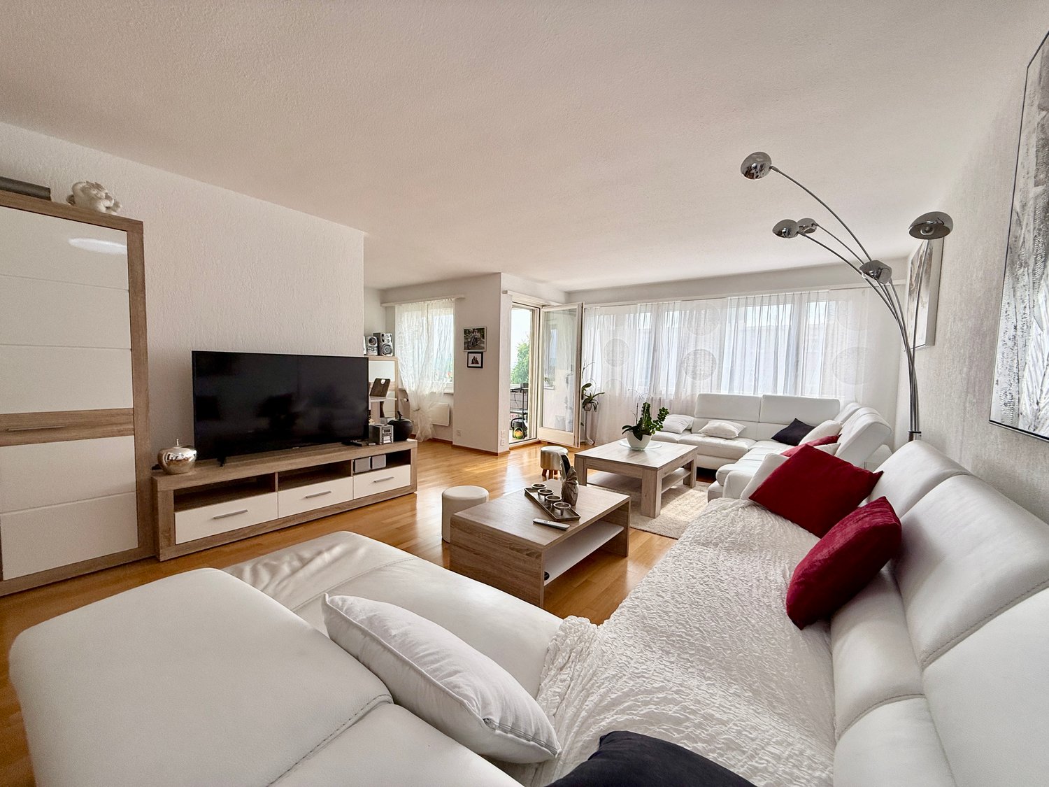 Spacious living room with large windows, white and gray furniture, wooden TV stand, coffee table, and floor lamp