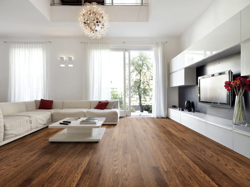 Spacious living room with hardwood flooring, large windows providing natural light, a white sectional sofa, a modern white coffee table, and a decorative chandelier overhead.
