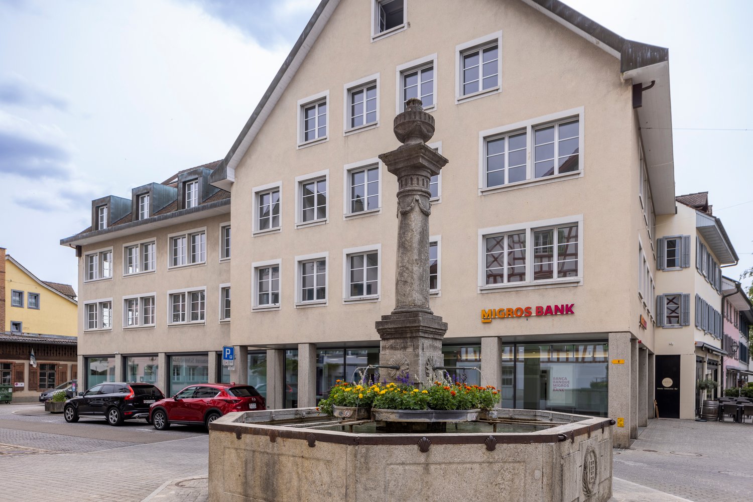 beige building with white windows, sign reads Micros Bank, parking area with 2 cars parked, fountain in front of building, table and chairs on sidewalk