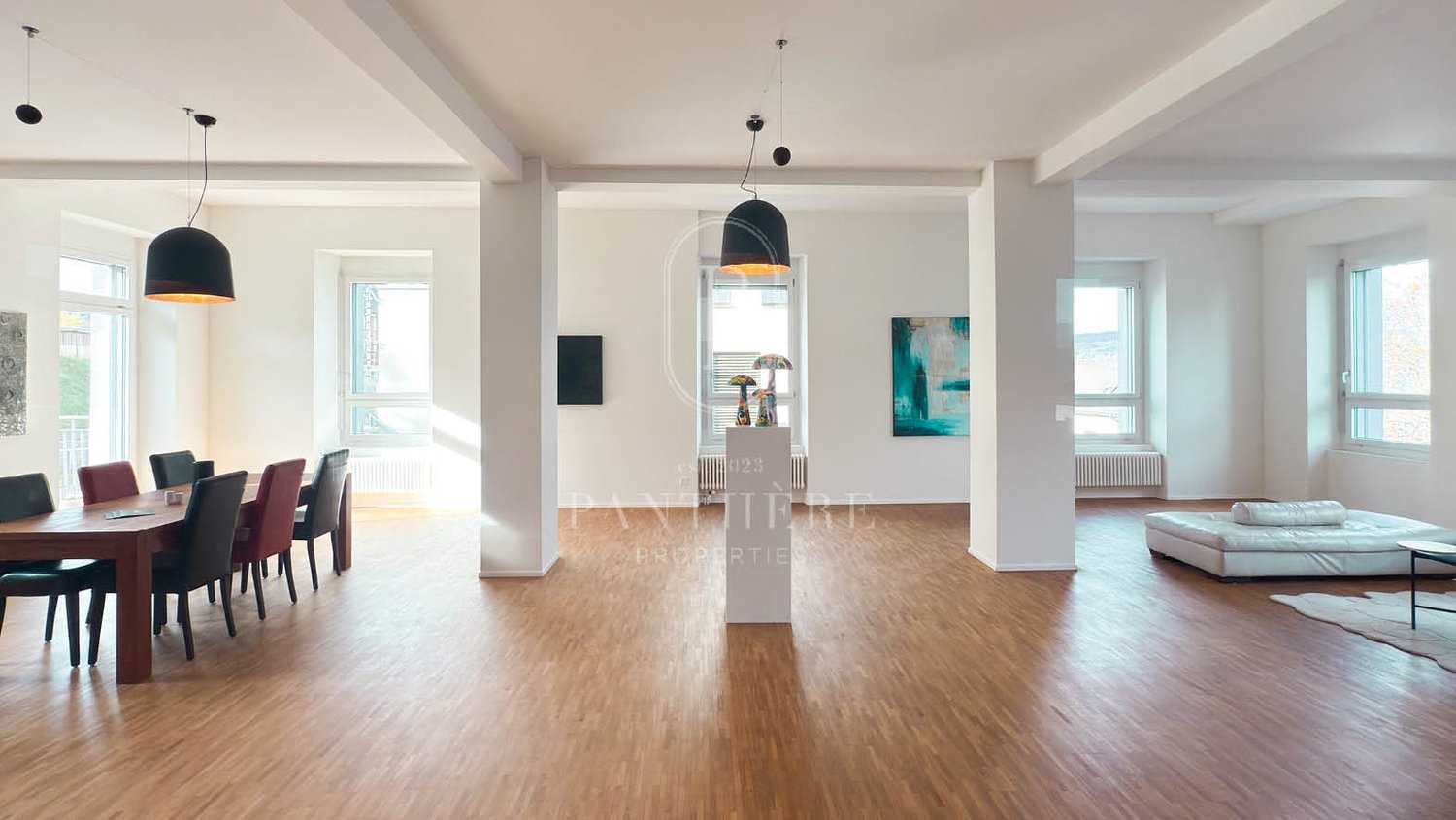 Open-plan room with large windows, wooden floor, minimalist decor, dining table and chairs, art pieces on the wall, radiator, and central pillars