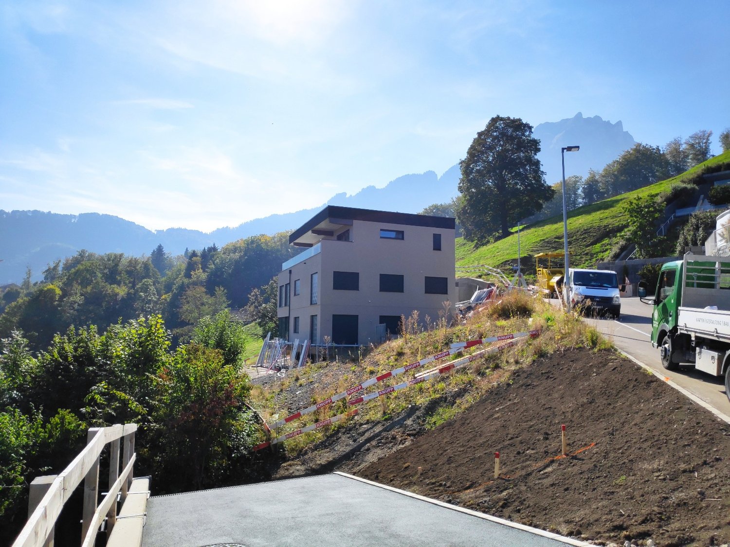 3-story modern residential building on a hillside, surrounded by trees and mountains. The building has a raised ground floor and is under construction, with construction equipment and materials visible on the site.