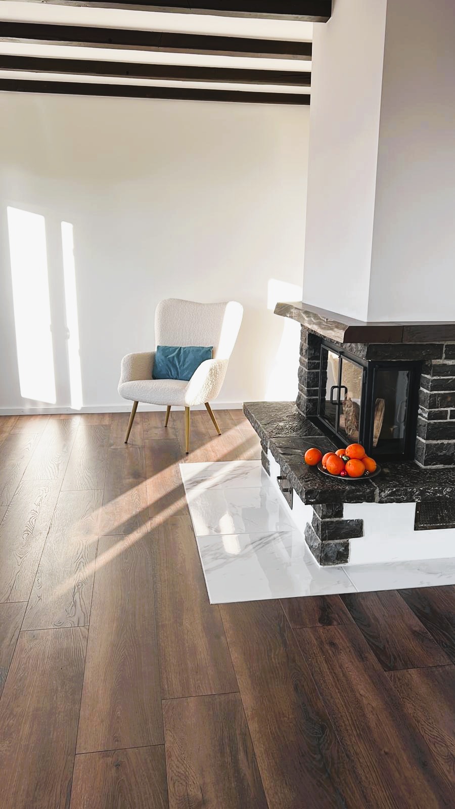Spacious living room with hardwood flooring, a white armchair, and a stone fireplace with a bowl of oranges on top.