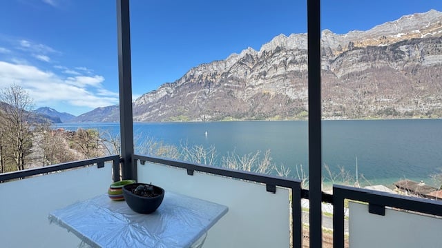 Balcony overlooking a lake surrounded by mountains, with a clear blue sky and a potted plant on a table