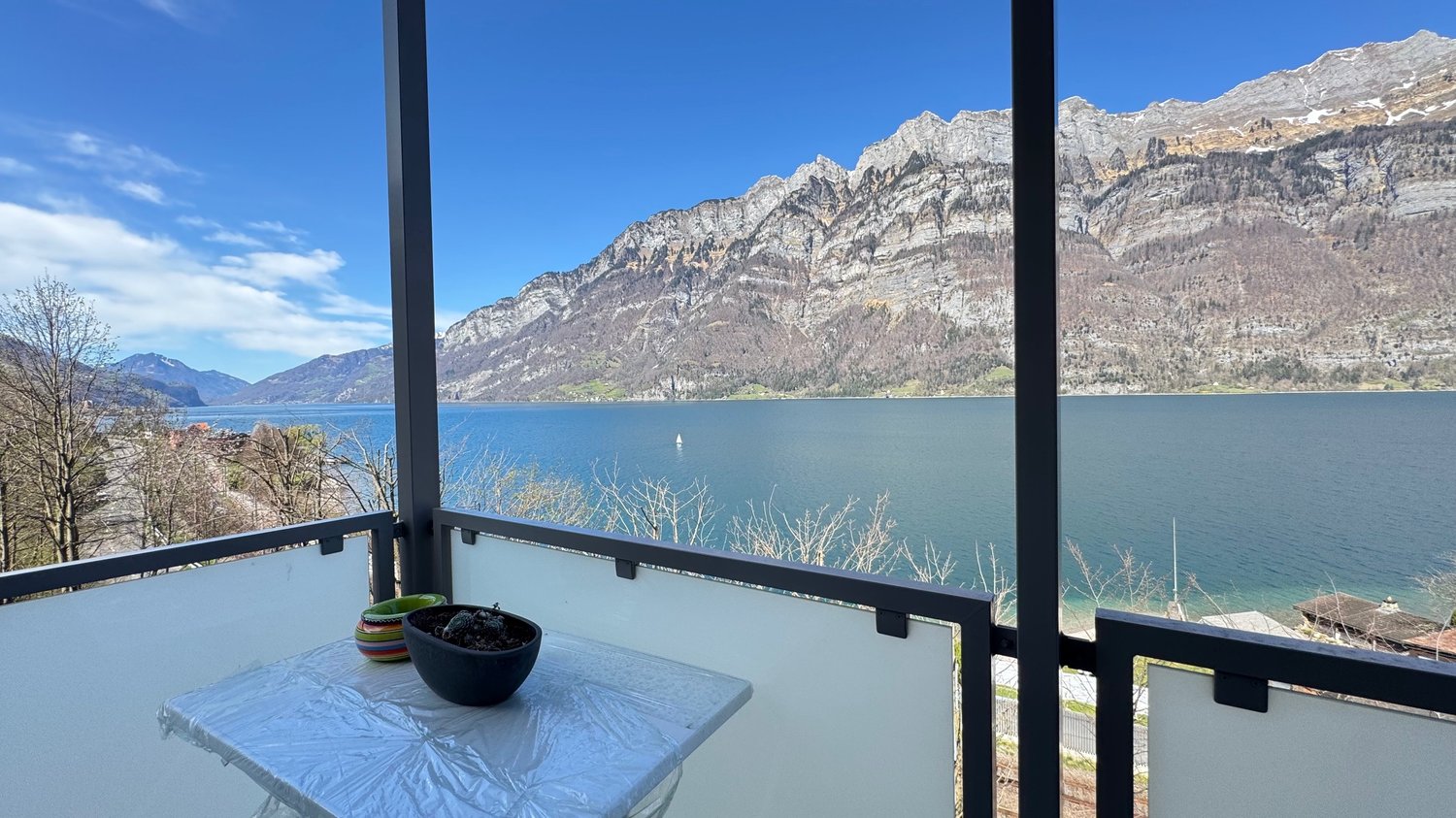 The image shows a balcony or terrace overlooking a scenic lake surrounded by snow-capped mountains. The balcony has a small table with a bowl of fruit, and the view through the glass railing is breathtaking, with the blue lake and distant peaks in the bac