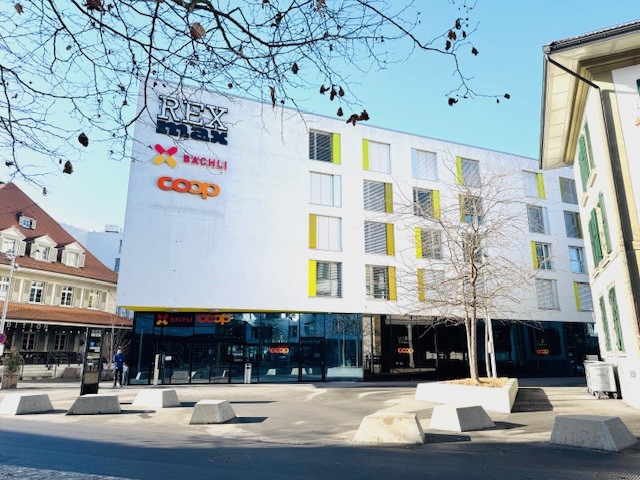 Modern white building with yellow accents, large glass windows, multiple floors, entrance with glass doors, signage for 'REX MAX' and 'COOP', concrete planters, street lights, and a tree in front.