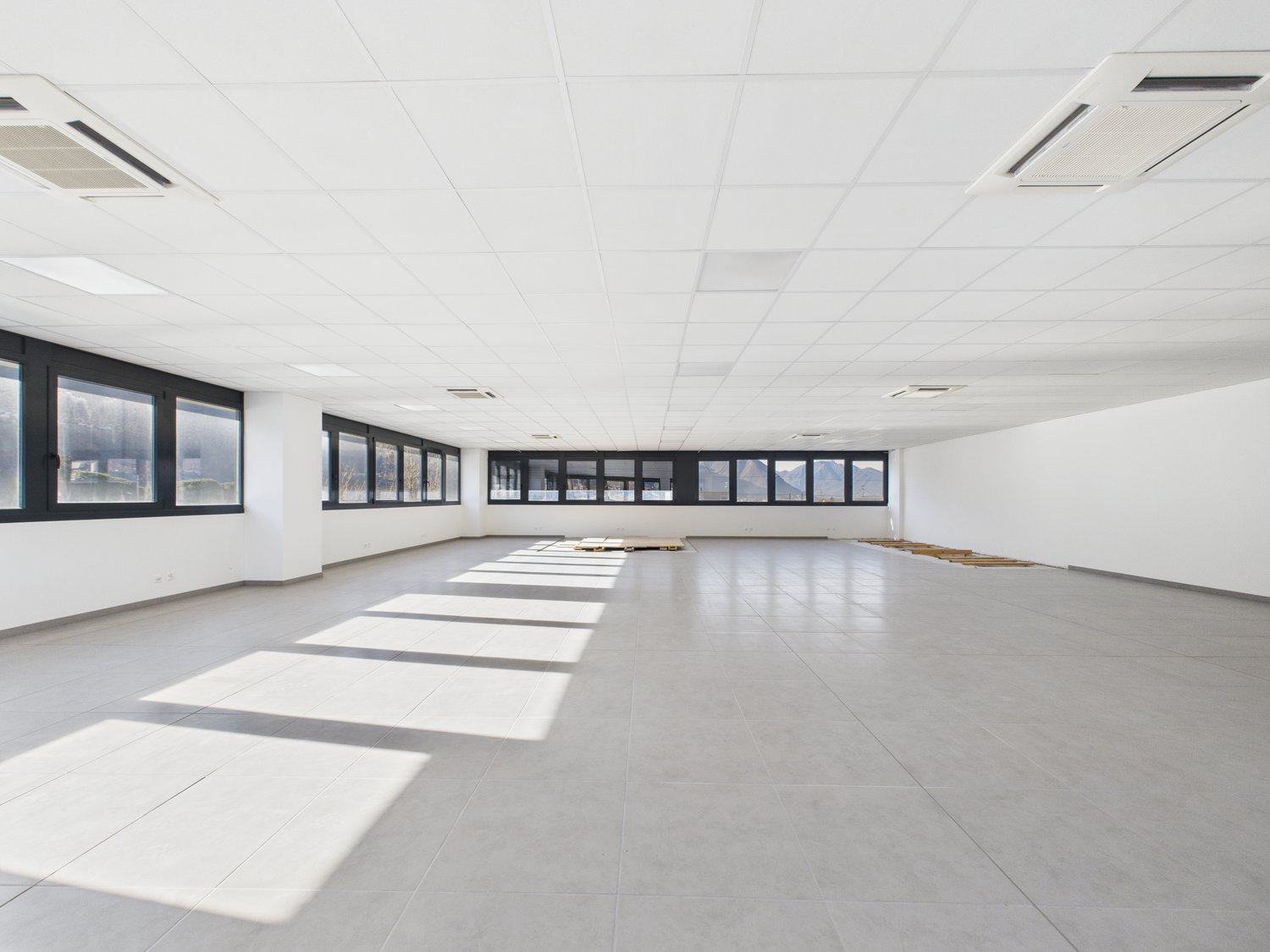 Empty room with white walls, gray tile flooring, large windows, and ceiling lights