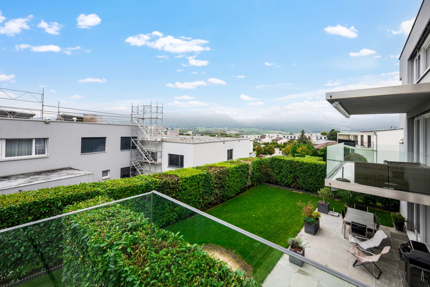 Outdoor terrace with a glass railing, well-kept hedges, and a seating area with chairs and a table, offering a view of the surrounding area.