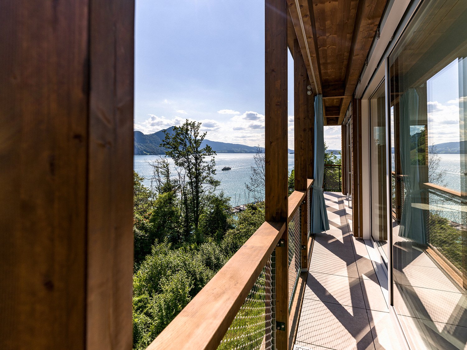 Wooden balcony with a panoramic view of a lake surrounded by mountains and trees. The balcony has large windows and a railing overlooking the scenic landscape.