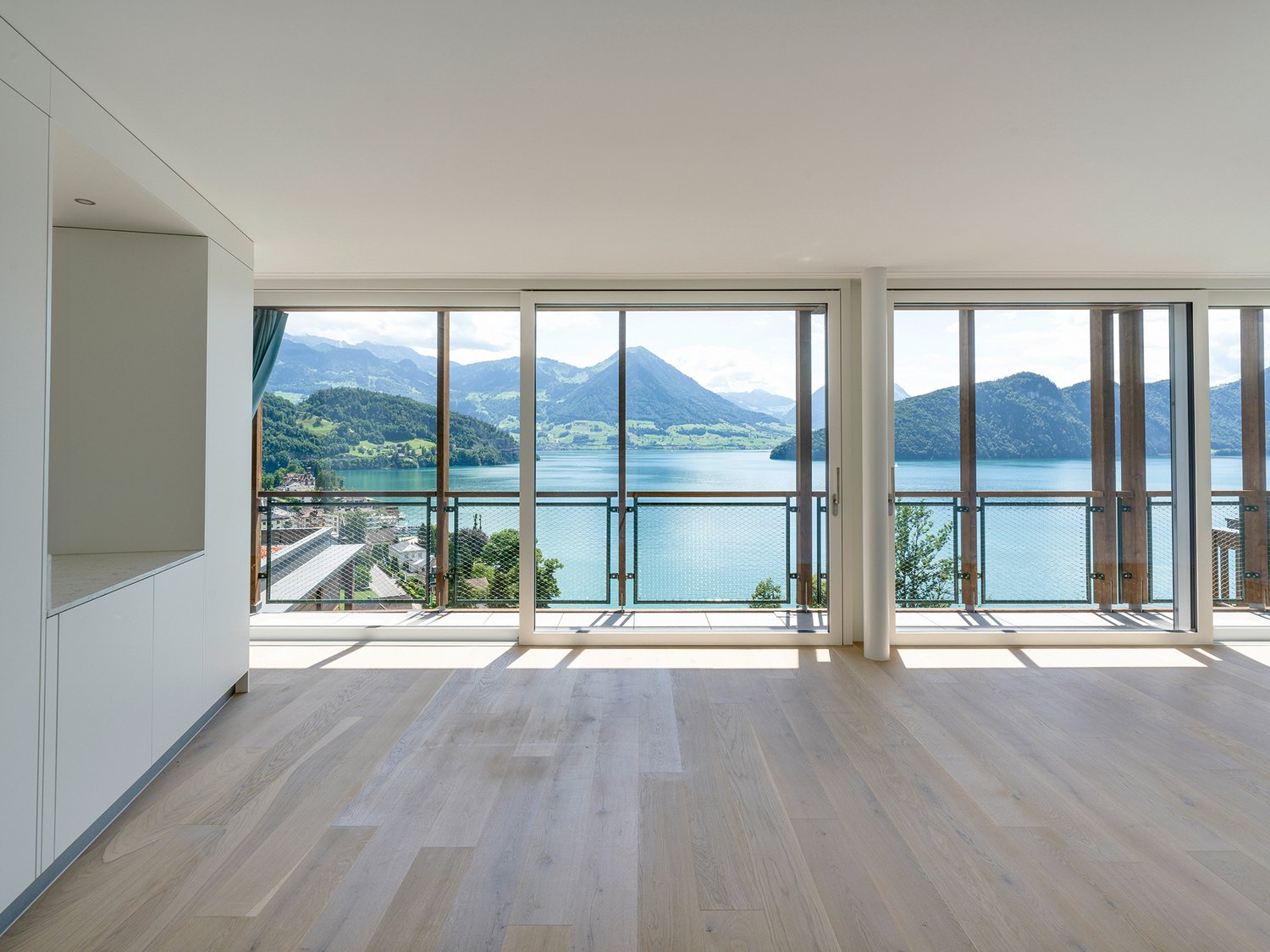 Spacious living room with large windows offering a panoramic view of a lake surrounded by mountains. The room has a minimalist design with light-colored hardwood floors and white walls. The windows provide abundant natural light and a serene, picturesque