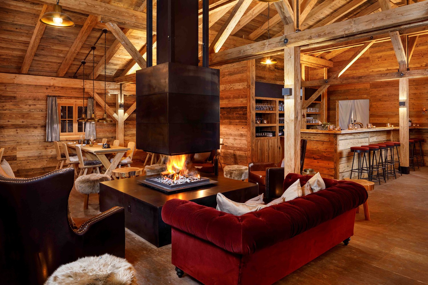 Rustic log cabin interior with exposed wooden beams and walls, a large fireplace with a fire burning, a red velvet sofa, a wooden dining table, and a bar area with shelves of glassware