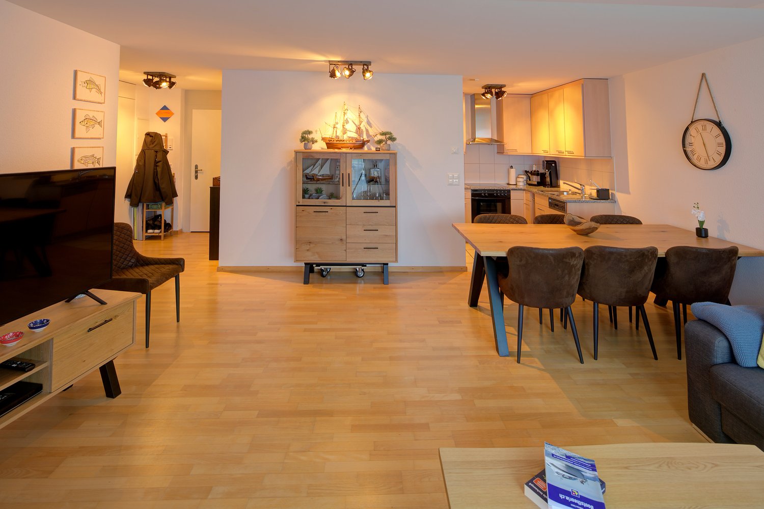 open living area with dining table and couch, wooden floor, large window, TV on stand, kitchen, cabinet, dining table with chairs
