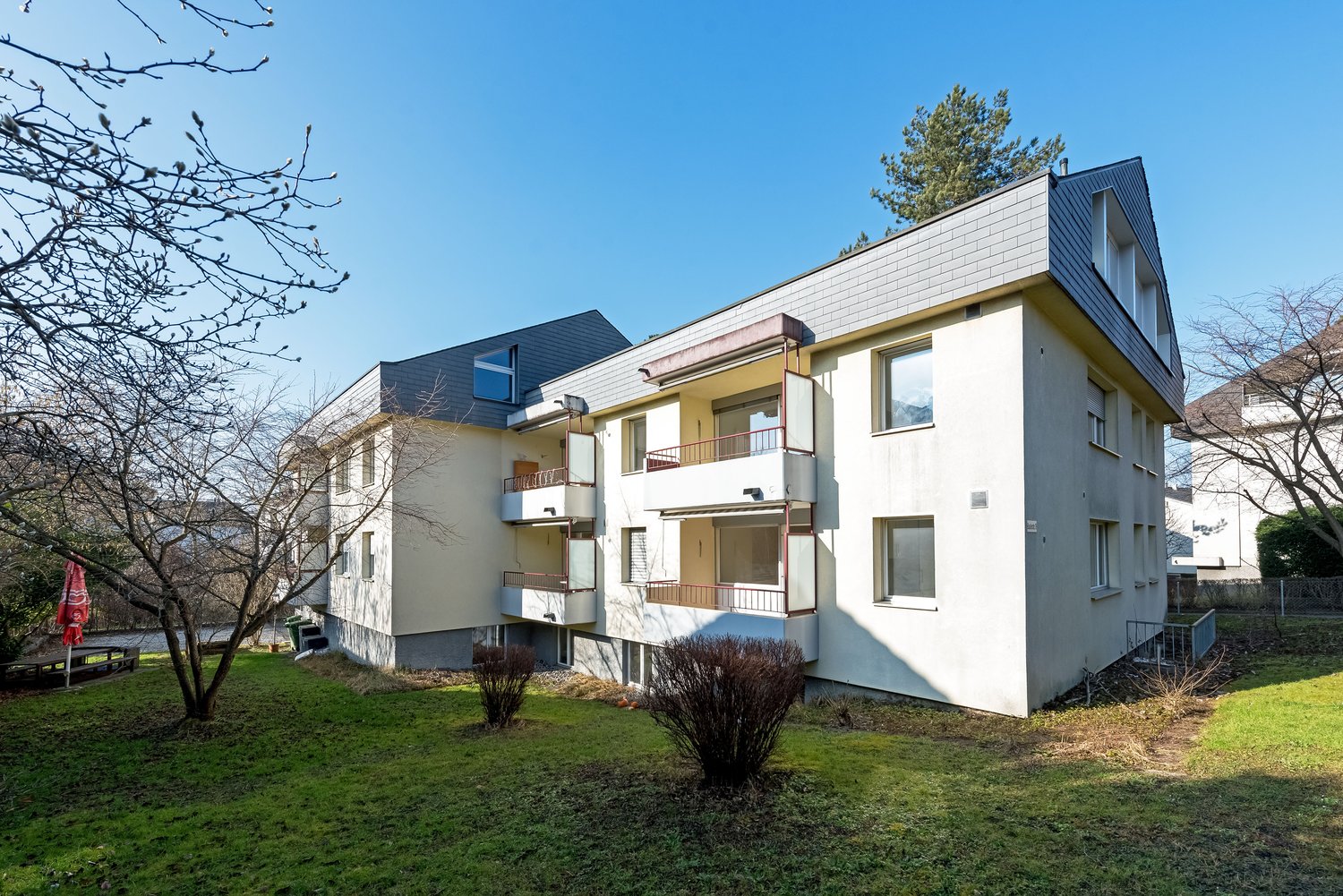 3-story apartment building with white walls, black roofing, and balconies. The building is surrounded by a grassy area with some trees and shrubs.