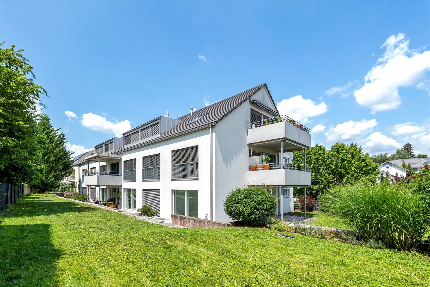 3-story apartment building with gray exterior, balconies, large windows, surrounded by lush green landscaping and trees