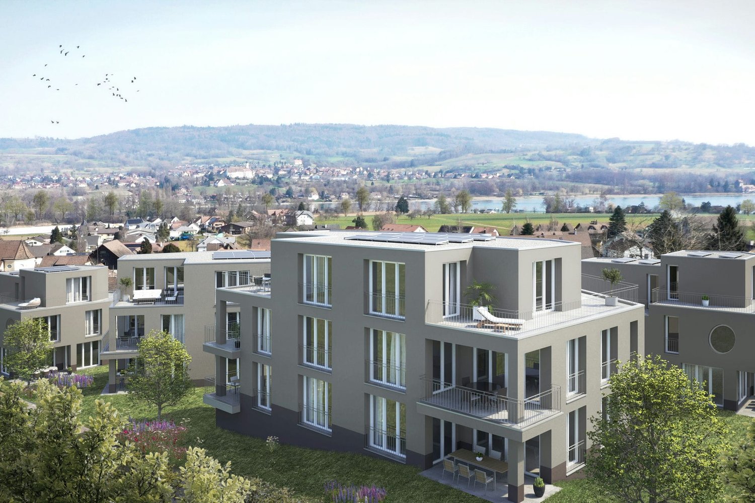 Multi-story apartment building with modern, gray exterior and large windows. The building has balconies on some units. The surrounding area includes a hilly, forested landscape with a small town or village in the distance.
