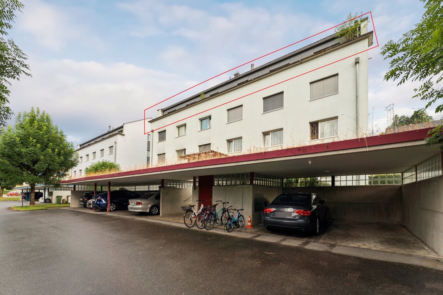 Multi-story apartment building with a covered parking area on the ground floor. The building has a red-colored roof and windows, and there are several cars and bicycles parked in the covered parking area.