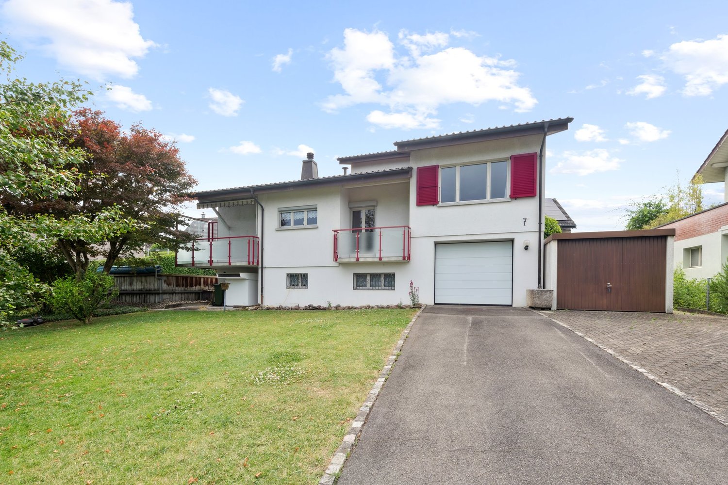 2-story house with white exterior, red shutters, balconies, garage, and a grassy yard with trees