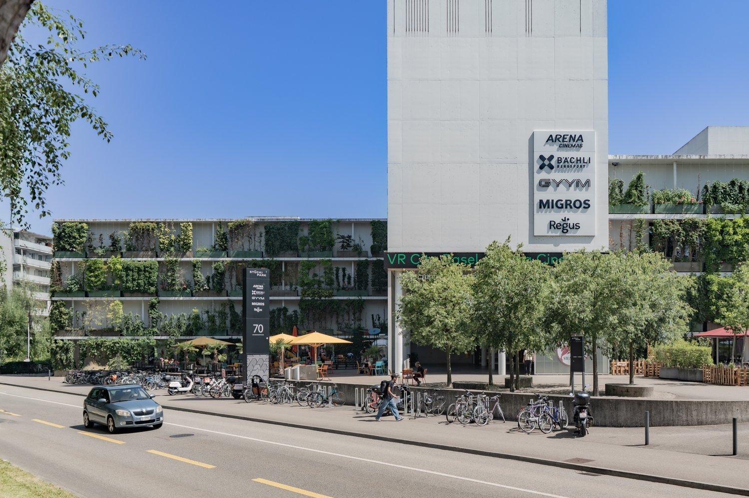 Modern commercial building with a vertical garden, outdoor seating, VR Cinema, arena cinemas, Bächli Bergsport, gym, Migros, Regus, sign boards on the front, large windows, multiple bicycles parked, trees, street, a moving car, road with yellow markings,